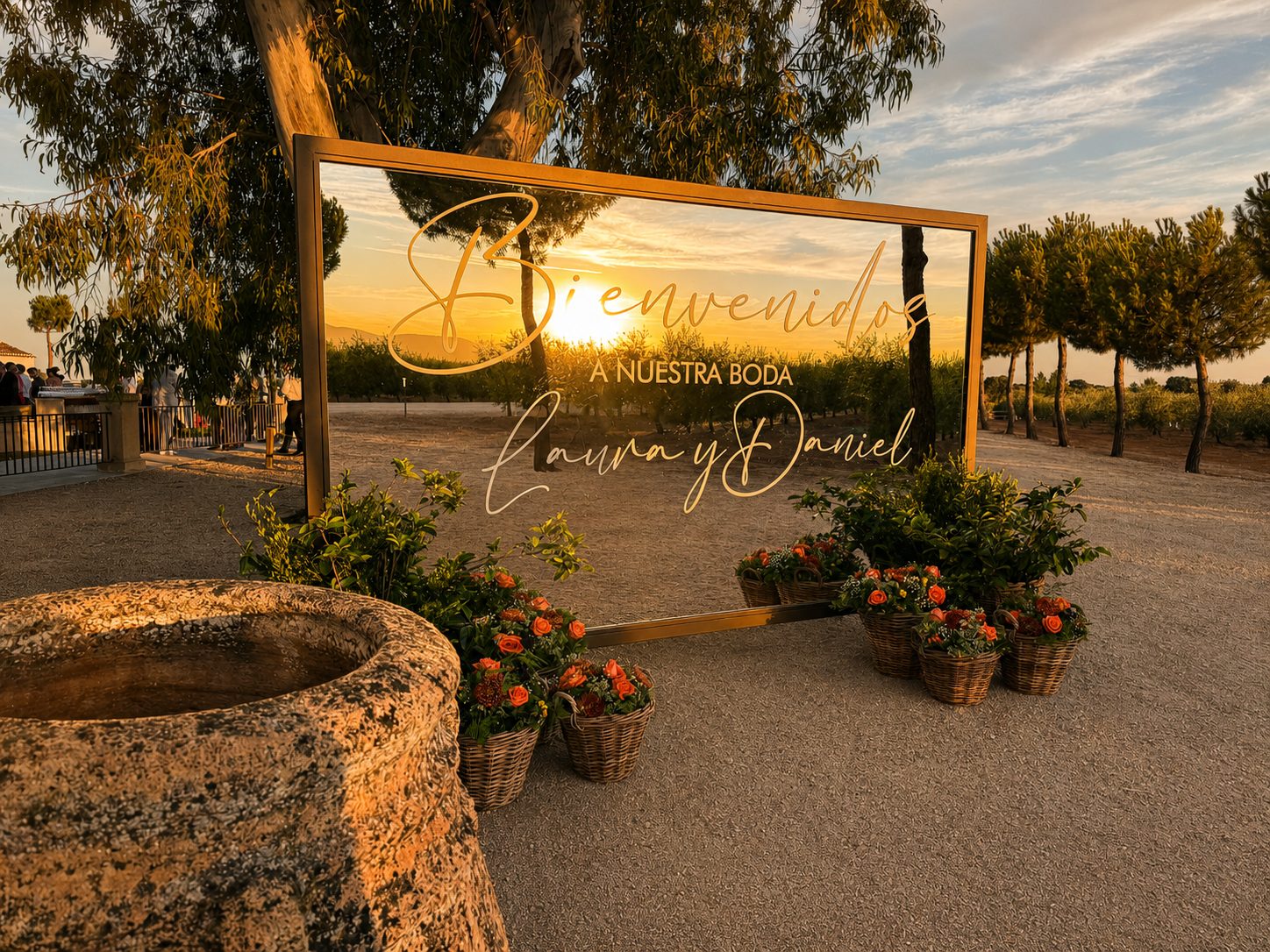 El espejo de bienvenida de la boda, rodeado de cestas de flores naranjas, refleja la puesta de sol sobre el olivar