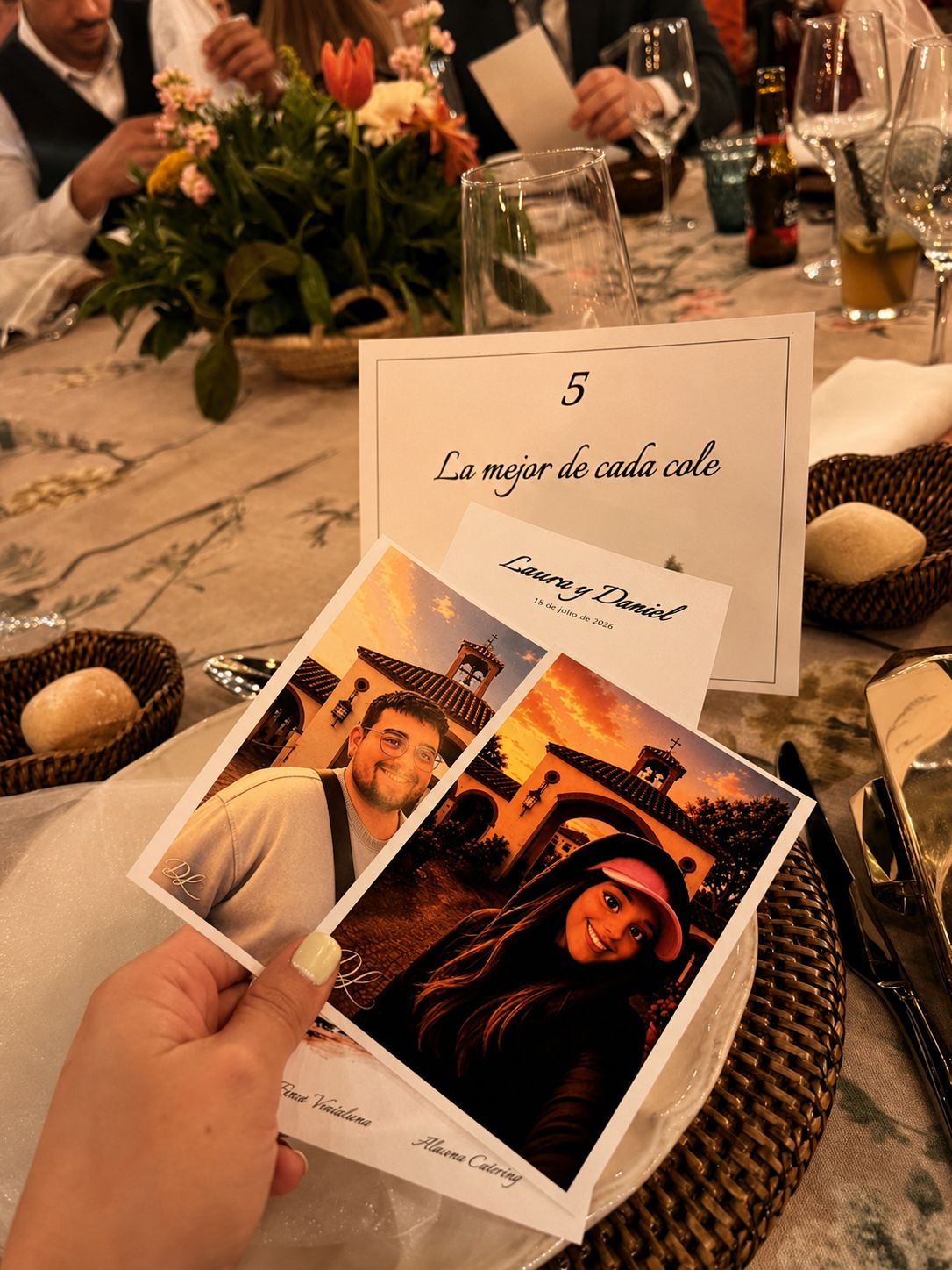 Una invitada sostiene la minuta ilustrada de la boda sobre la mesa vestida, con el número y el nombre de la mesa detrás