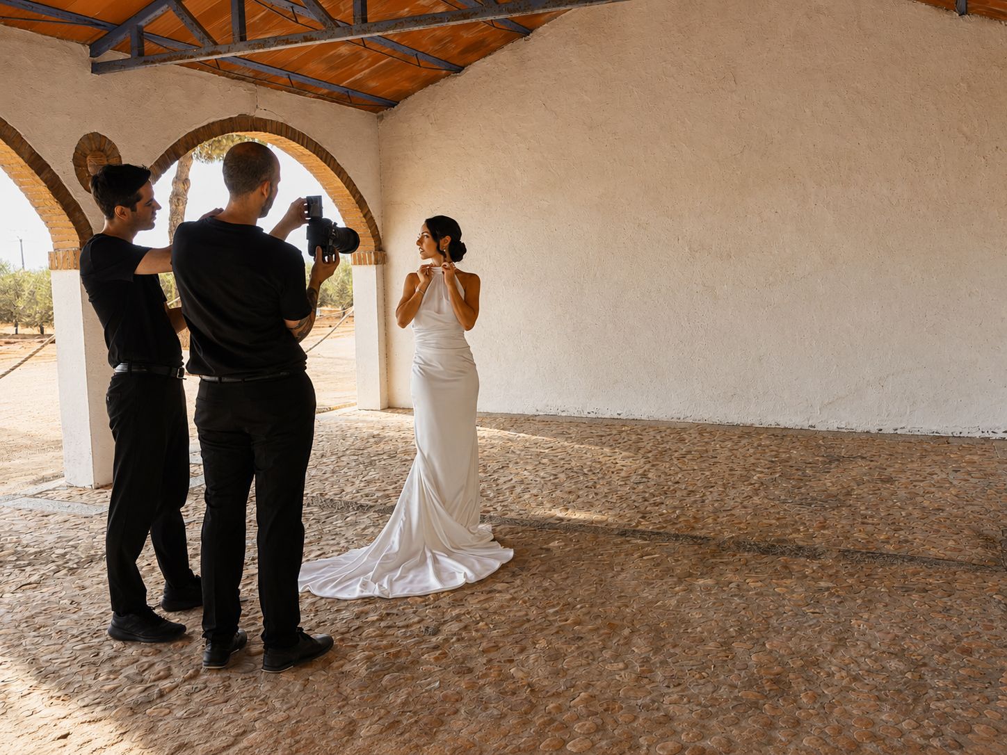 La novia se ajusta el vestido de raso mientras el fotógrafo la retrata bajo la arcada de la finca