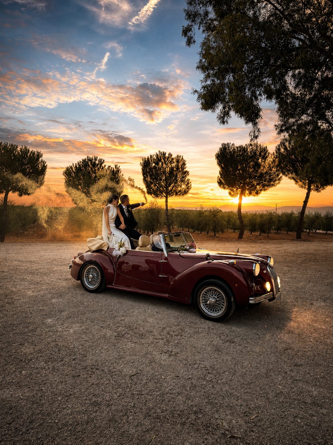 Los novios, sentados sobre un descapotable clásico granate, contemplan el atardecer sobre el olivar con una bengala de humo