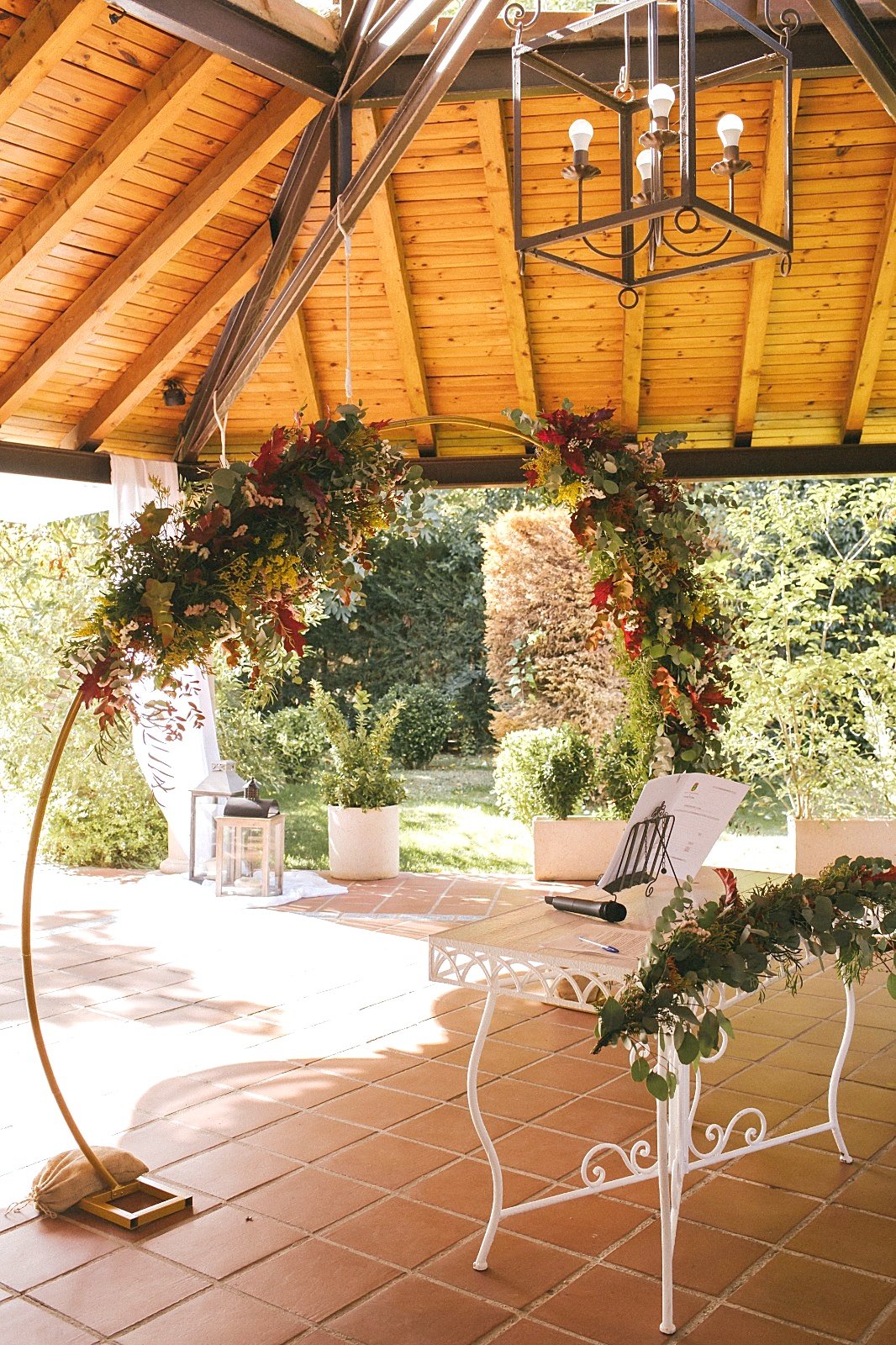 El altar de la ceremonia: un arco de flores rojas y hojas de otoño sobre una mesa blanca, con el jardín al fondo