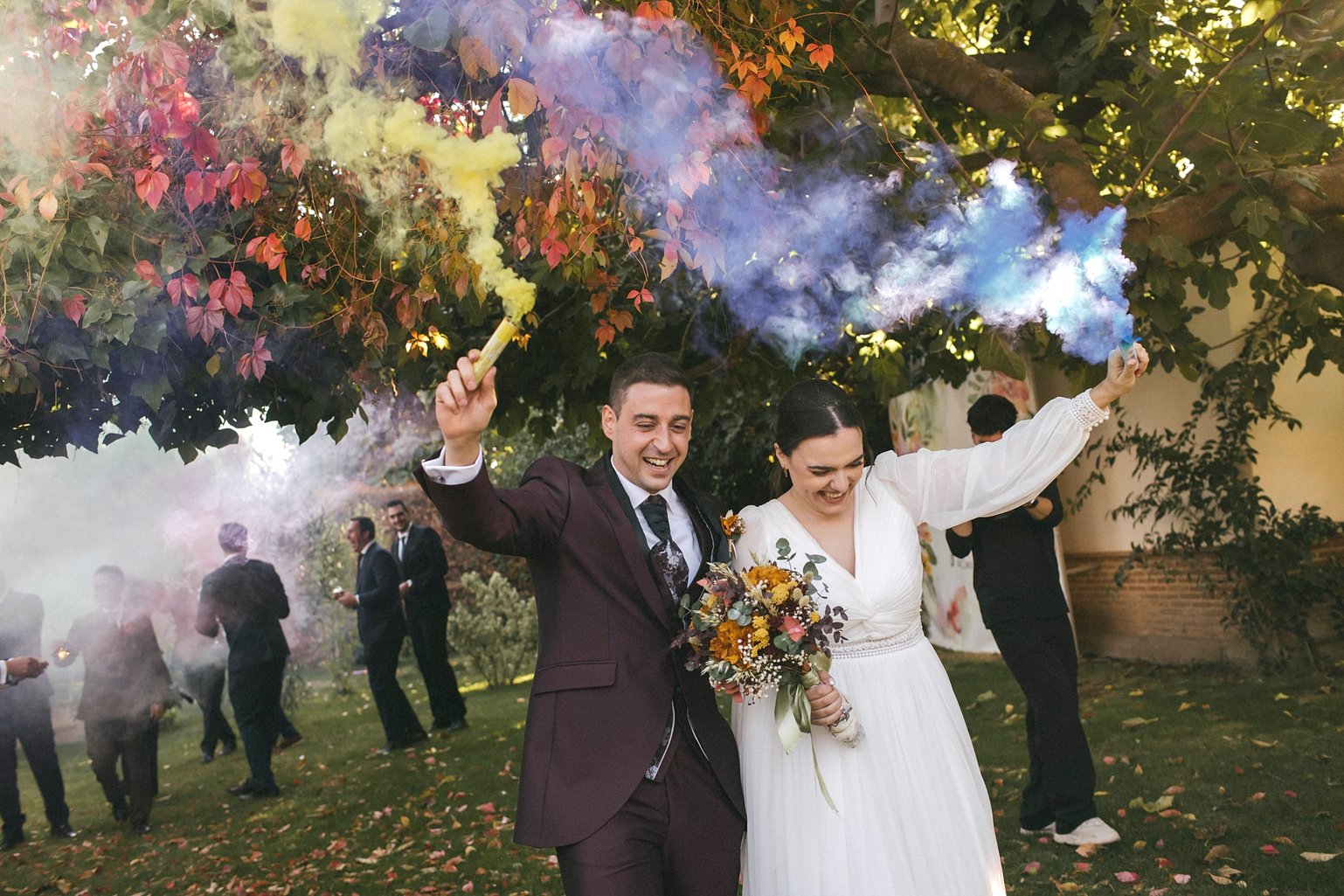 Los novios ríen bajo un árbol de hojas otoñales mientras sostienen bengalas de humo de colores