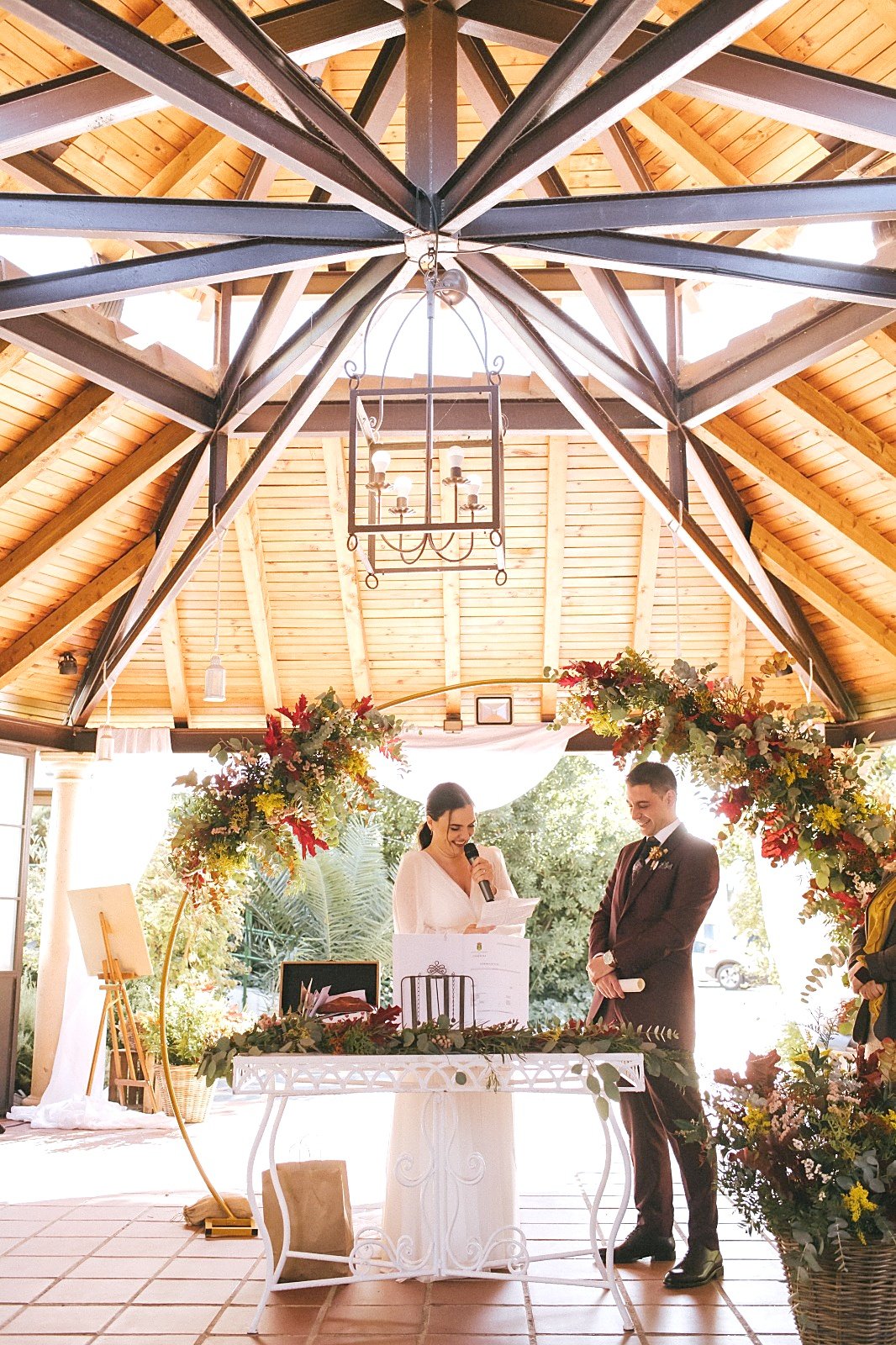La novia lee sus votos con un micrófono junto al novio, bajo un cenador de madera con un arco de flores de otoño