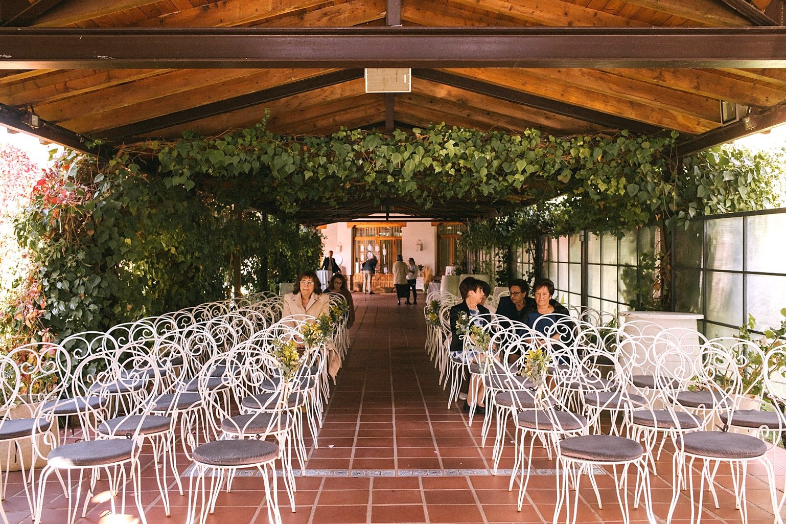 El pasillo de la ceremonia bajo una pérgola cubierta de hiedra, con sillas blancas de forja y ramos a los lados