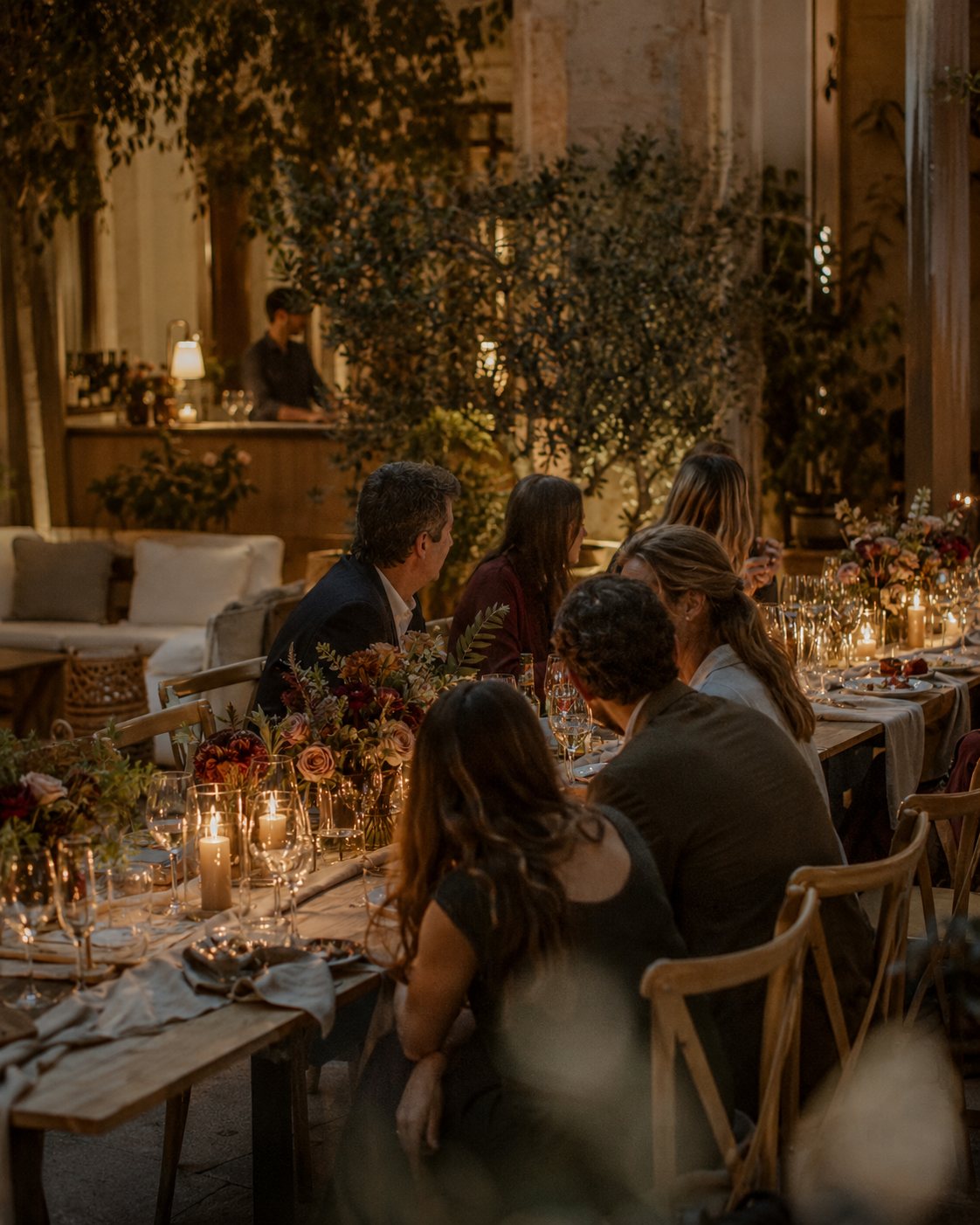 Invitados sentados a una mesa larga iluminada con velas, conversando durante la cena