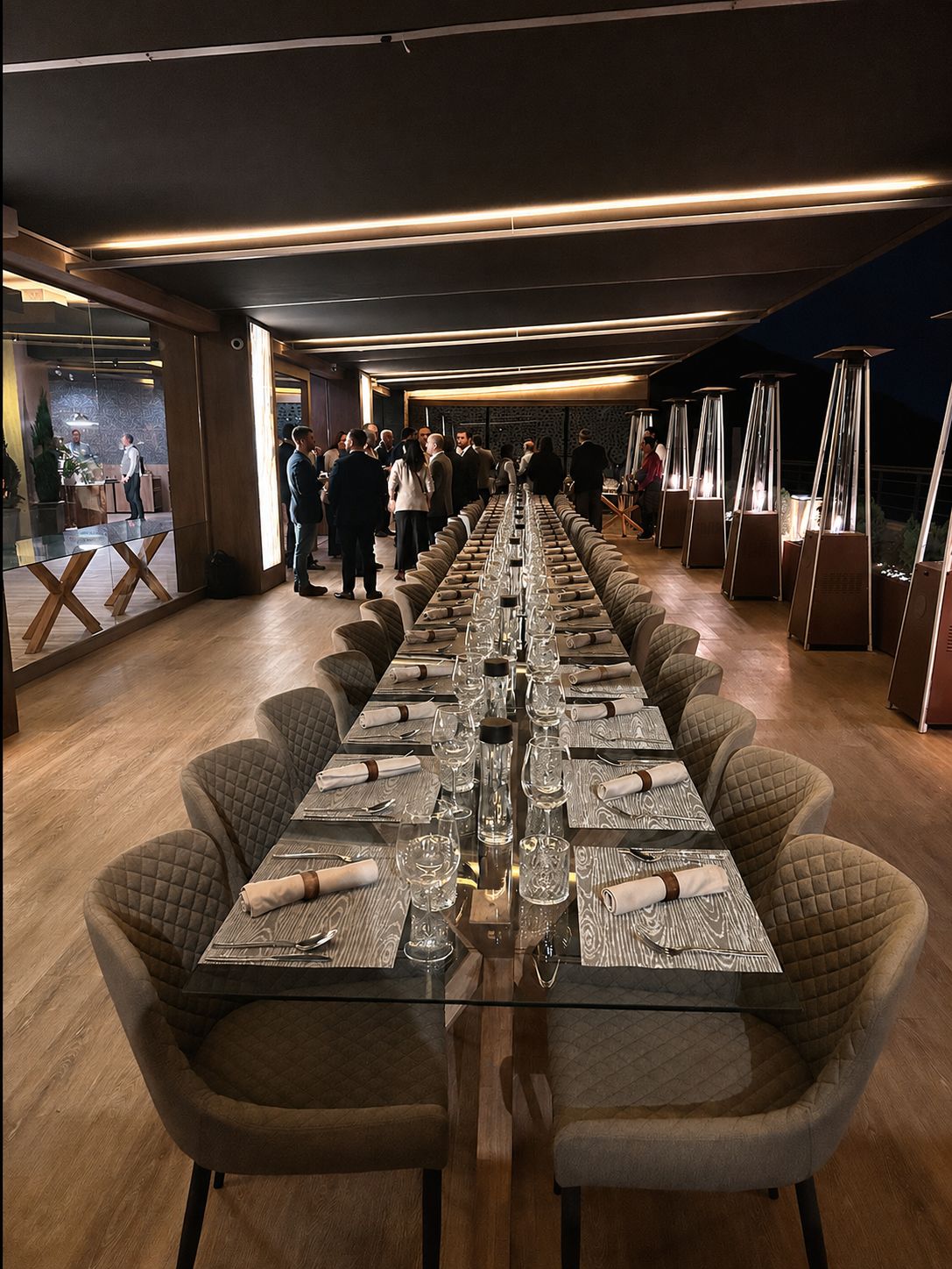 Larga mesa vestida para una cena de empresa en una terraza cubierta con vistas, con los invitados charlando de fondo al anochecer